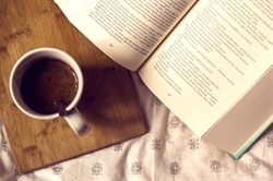 [ai] A white mug with dark liquid resting on a wooden board next to an open book with text pages, set on a patterned fabric surface.